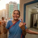 A middle-aged Indian woman in a saree holds a steel key and is smiling as she opens the door of a modern flat. Behind her, other family members look excited